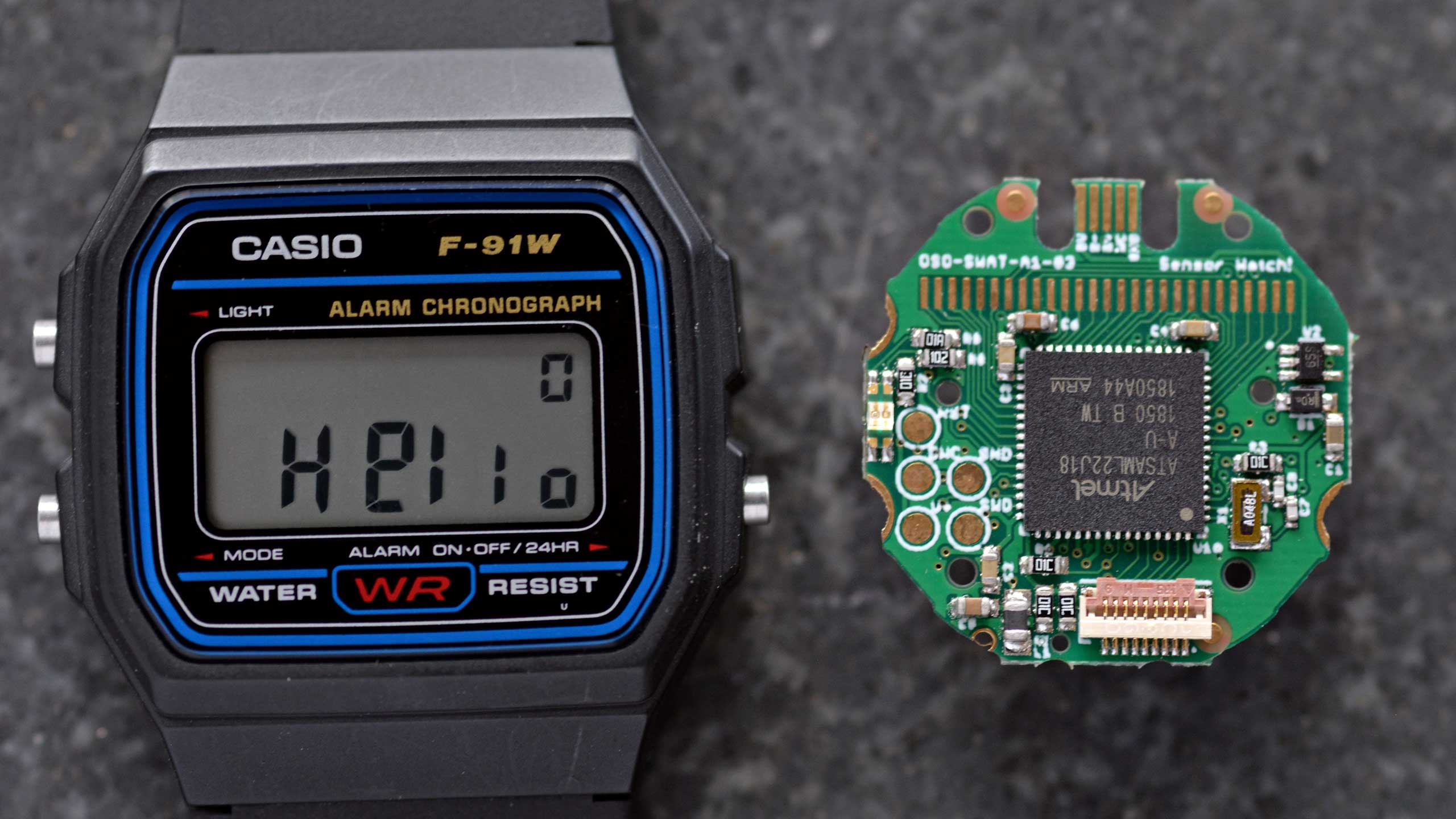 Photo: a close-up of the Sensor Watch circuit board, and a wristwatch displaying the word “Hello”